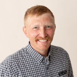 Professional headshot of a smiling man with short blond hair and a mustache, wearing a blue and white checkered button-down shirt against a light neutral background.