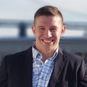 Photo of a man with short brown hair standing in front of a bridge with a button down shirt and suit jacket.