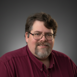Professional headshot of a smiling man with dark hair, glasses, and a gray goatee, wearing a burgundy button-down shirt against a gray gradient background.