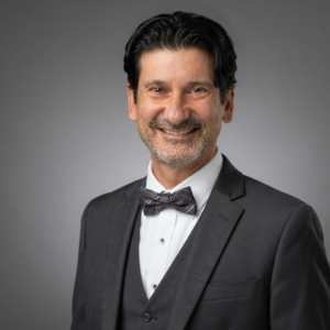 Professional headshot of a smiling man with dark hair and a salt-and-pepper beard, wearing a charcoal suit, vest, white shirt, and bow tie against a gray background.